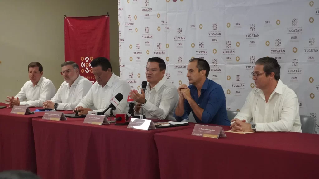 Seis hombres sentados en una mesa con manteles rojos en conferencia de prensa, fondo con logos del Gobierno de Yucatán