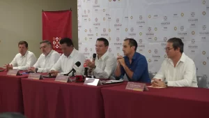 Seis hombres sentados en una mesa con manteles rojos en conferencia de prensa, fondo con logos del Gobierno de Yucatán