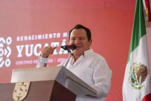 Hombre con guayabera blanca hablando en podio con micrófono frente a fondo rojo y bandera de México