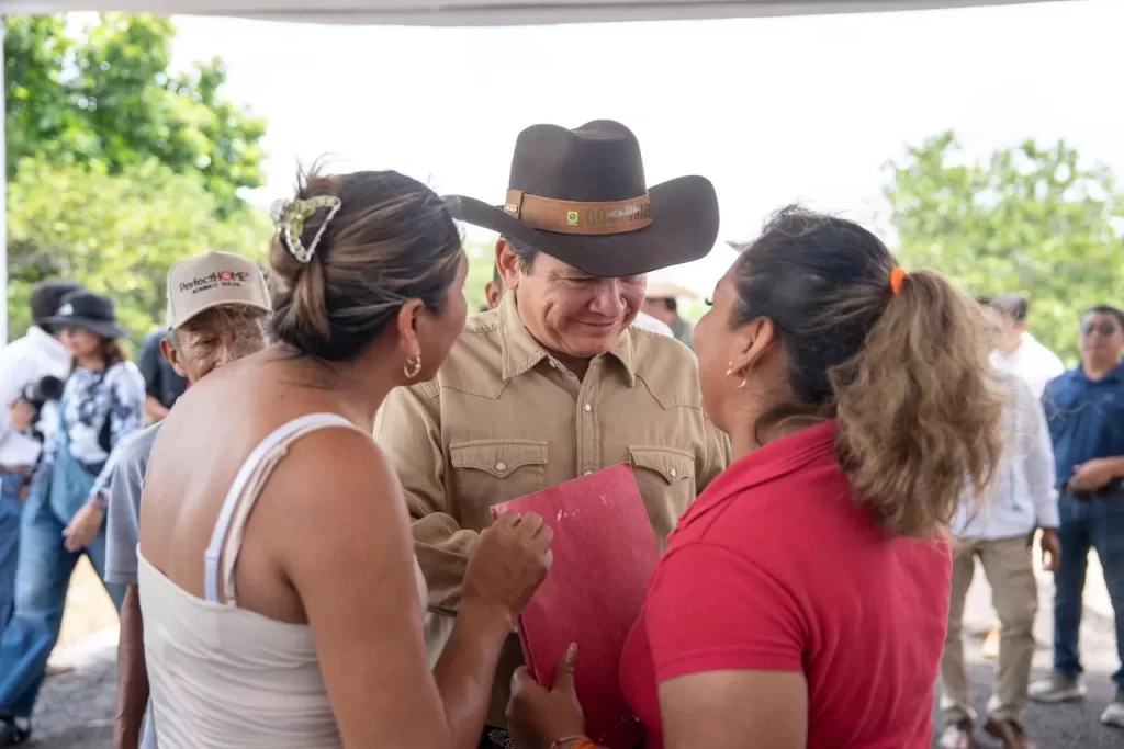 Hombre con sombrero marrón conversa con dos mujeres en un evento al aire libre