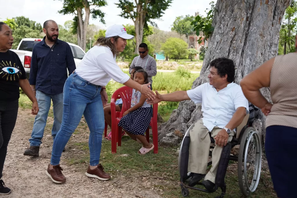Mujer con gorra blanca saludando de mano a hombre en silla de ruedas bajo un árbol