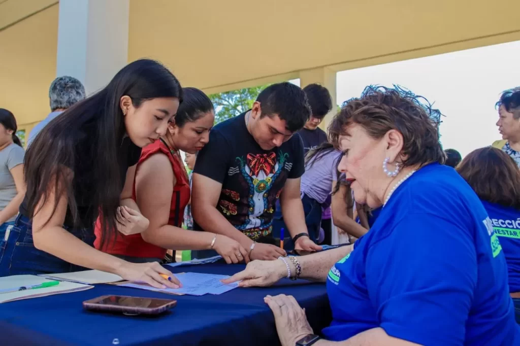 Personas jóvenes llenando formatos en mesa con ayuda de mujer adulta en evento social