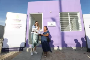 Dos mujeres sonrientes frente a casa morada sosteniendo letrero vivienda digna