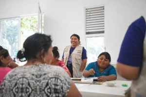 Varias mujeres participando en un taller de pintura en un espacio cerrado, una de ellas sonríe y lleva chaleco blanco