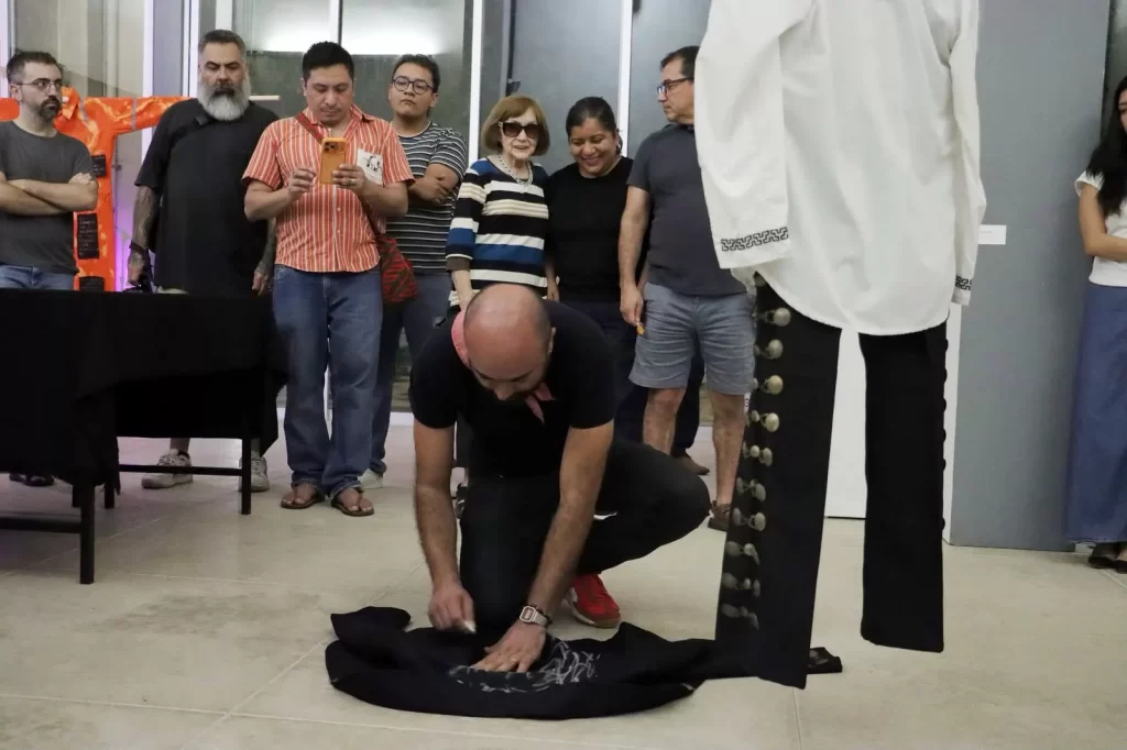 Hombre calvo agachado pintando una prenda negra en el suelo mientras un grupo de personas observa en un espacio cerrado