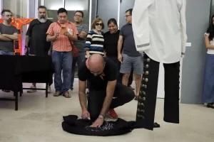 Hombre calvo agachado pintando una prenda negra en el suelo mientras un grupo de personas observa en un espacio cerrado