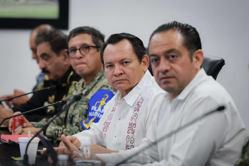 Grupo de cinco hombres en reunión, algunos con uniformes militares y otros con guayaberas blancas bordadas, sentados frente a