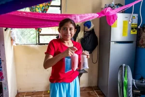 Niña sonriente con camiseta roja y falda azul sostiene bolsas de plástico con agua y colorante dentro de una casa