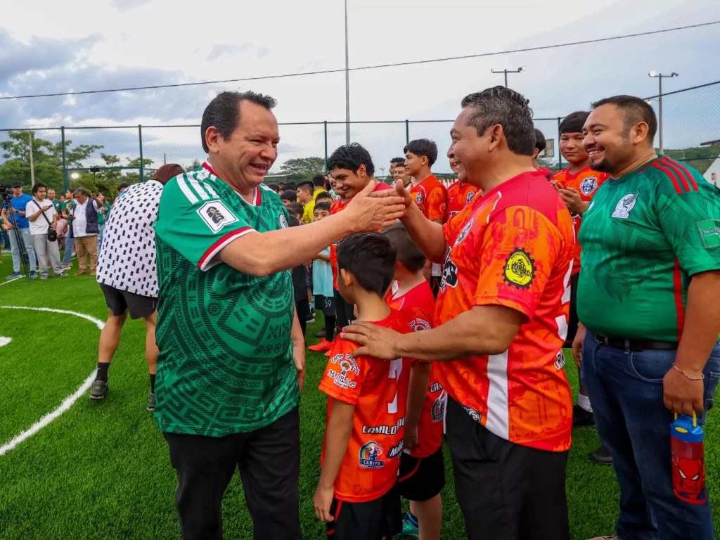 Hombres saludándose en cancha de futbol con niños y personas al fondo