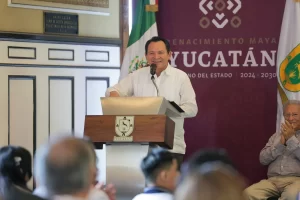 Gobernador de Yucatán dando un discurso en podium con bandera y escudo del estado al fondo