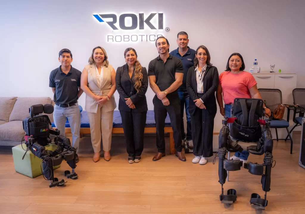 Siete personas de pie en oficina con logo ROKI Robotics y dos exoesqueletos frente a ellos