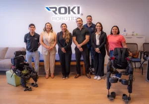 Siete personas de pie en oficina con logo ROKI Robotics y dos exoesqueletos frente a ellos