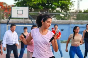 Mujer joven con camiseta rosa y diadema sosteniendo micrófono y bailando en cancha deportiva con varias personas atrás