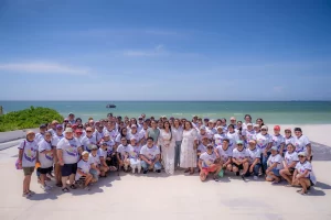 Gran grupo de personas posando en la playa frente al mar con camiseta blanca y gorras
