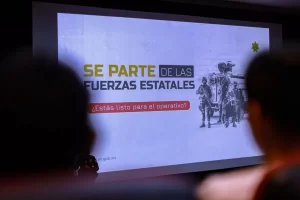 Proyección de pantalla con texto de convocatoria a las fuerzas estatales y dibujo de militares con vehículo blindado