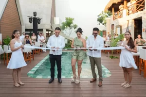 Tres personas cortan una cinta blanca en ceremonia frente a una alberca con dos mujeres vestidas de blanco sosteniendo la cin