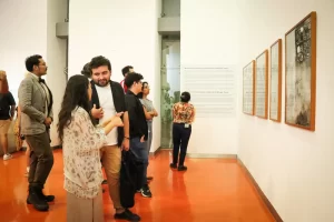 Varias personas de pie observando cuadros y texto en la pared de una galería de arte con piso naranja