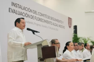 Hombre con guayabera blanca hablando en podio en evento oficial sobre evaluación de floración de algas nocivas en Yucatán