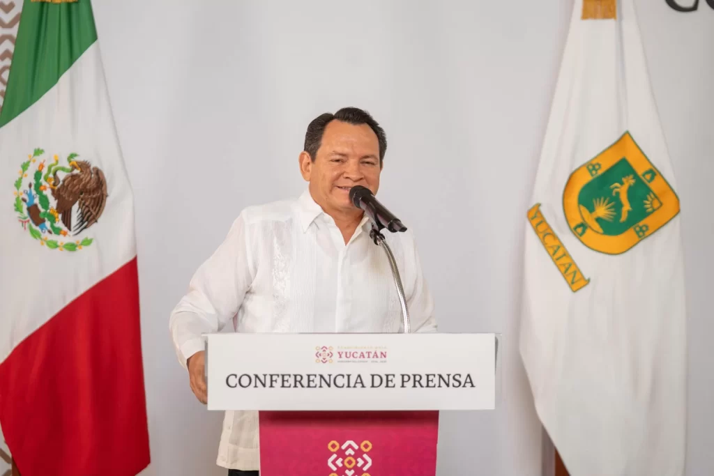 Hombre dando conferencia de prensa con bandera de México y Yucatán al fondo