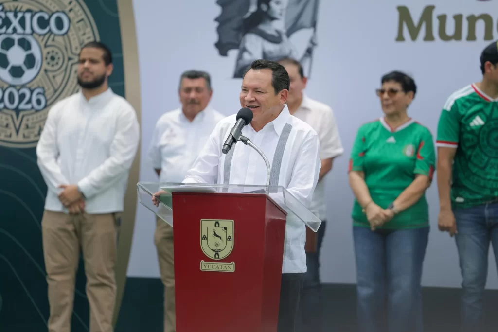 Hombre hablando en podio con logo de Yucatán, con grupo de personas detrás, fondo con logo México 2026