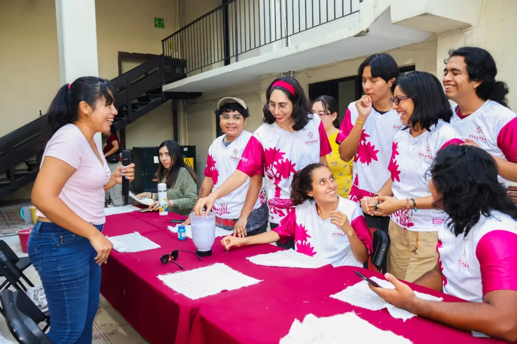 Grupo de jóvenes con camisetas blancas y rosas participando en actividad con licuadora sobre mesa con mantel rojo