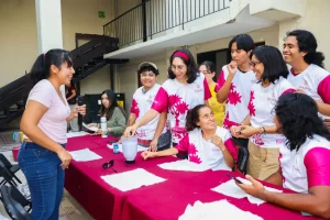 Grupo de jóvenes con camisetas blancas y rosas participando en actividad con licuadora sobre mesa con mantel rojo