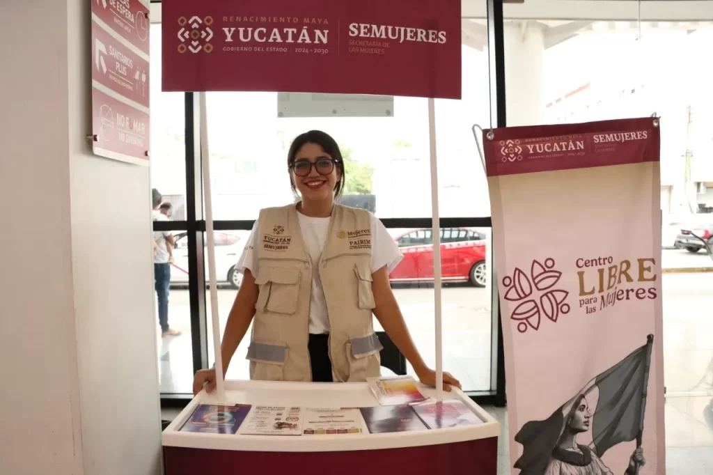 Joven mujer con gafas sonriendo en un módulo informativo de SEMUJERES y Gobierno de Yucatán
