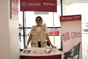 Joven mujer con gafas sonriendo en un módulo informativo de SEMUJERES y Gobierno de Yucatán