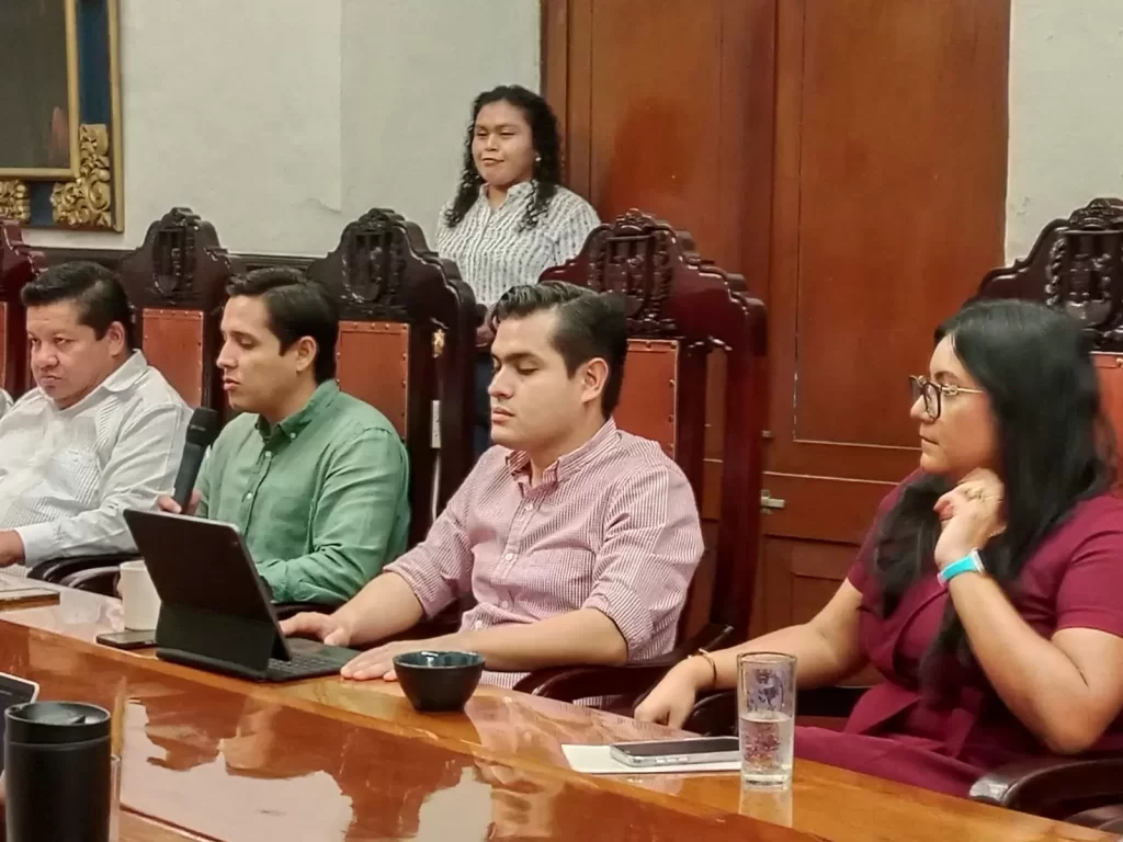 Cinco personas sentadas en una mesa de madera en una sala con sillas de respaldo alto, una persona usando micrófono y laptop