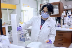 Persona con bata blanca, guantes y cubrebocas trabajando en laboratorio de química