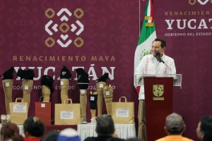 Persona dando un discurso en podio con bandera mexicana y piercings ortopédicos expuestos detrás