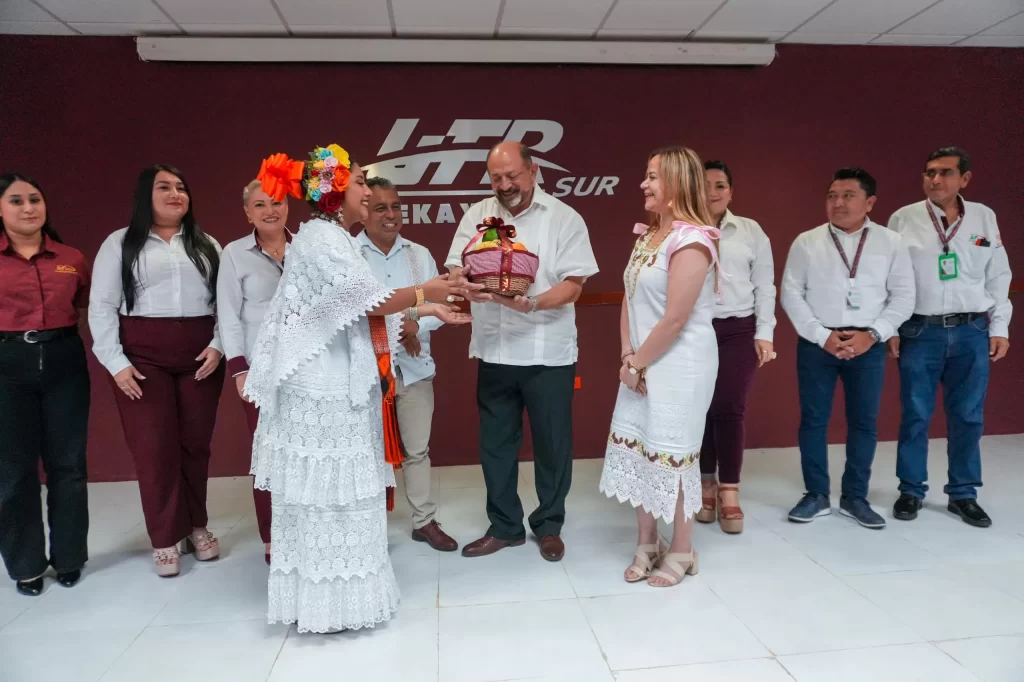 Persona con vestido tradicional entregando obsequio a un hombre con guayabera blanca en grupo de personas en interior