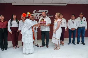 Persona con vestido tradicional entregando obsequio a un hombre con guayabera blanca en grupo de personas en interior