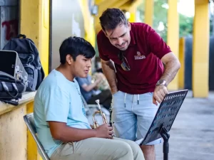 Profesor mostrando partituras a alumno con trompeta en espacio abierto con columnas amarillas