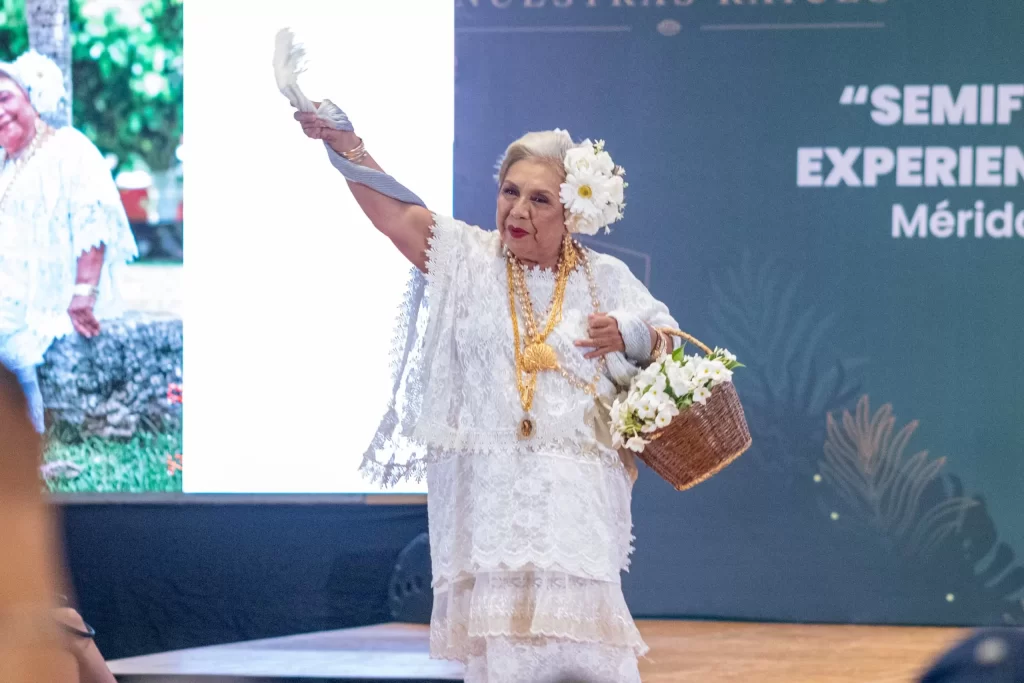 Mujer mayor con vestido blanco de encaje y flores en la cabeza, con collar dorado y canasta con flores, en un escenario