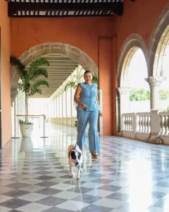 Mujer con conjunto azul caminando con perro blanco con manchas negras en corredor con piso ajedrezado y arcos de piedra