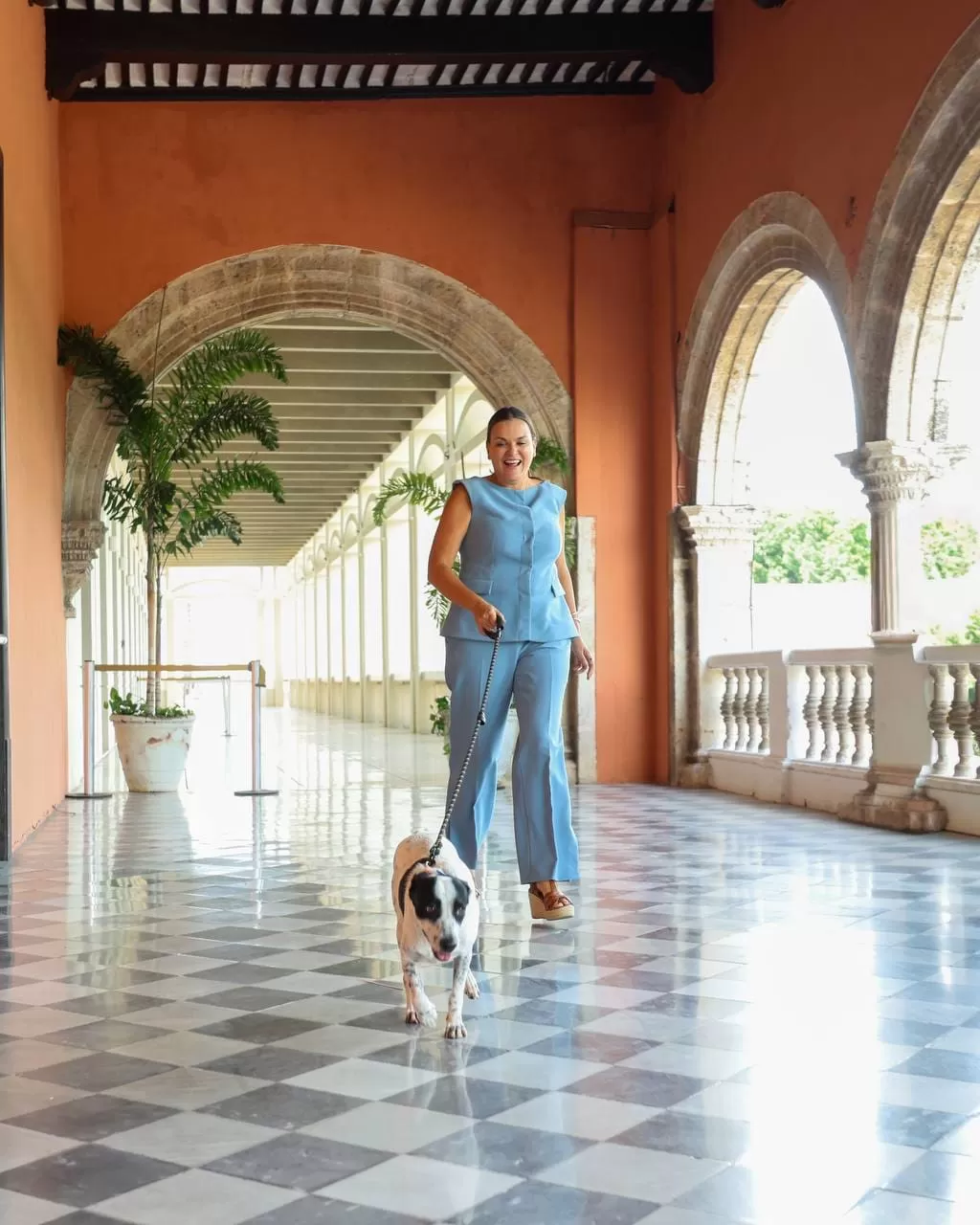 Mujer con conjunto azul caminando con perro blanco con manchas negras en corredor con piso ajedrezado y arcos de piedra